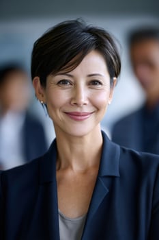 Confident woman smiles in business attire, with blurred colleagues behind her photo