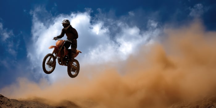 Motorcycle rider jumps on dirt terrain under blue sky with clouds and blowing dust photo