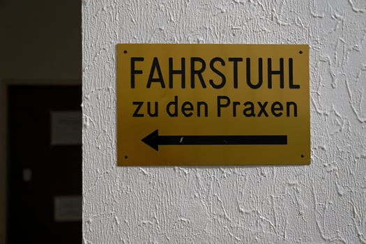 Gold colored sign mounted with four screws on uneven white painted wall showing German text Fahrstuhl zu den Praxen with left pointing arrow photo
