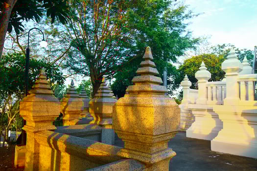 A Thai temple balcony glows at sunset, casting long shadows along the stone railing. Lush trees and garden surround the ancient architecture, evoking peace and heritage. photo