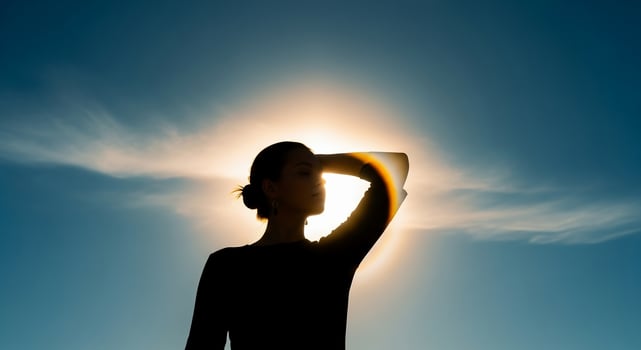 Silhouette of a woman shielding her eyes from the sun against a bright sky with clouds photo
