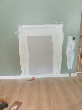 Wall repair with patching and sanding process in progress, showing partially painted wall with rectangular patch and smaller patch on right side, in home interior photo