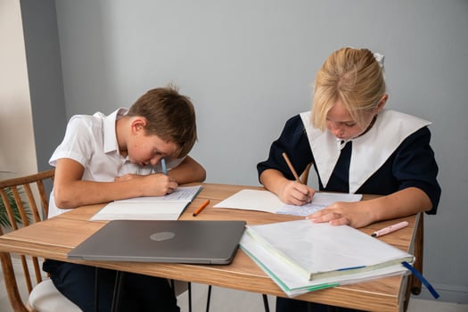 Children School Homework Studying - Two children are sitting at a table and writing in their notebooks. A laptop is on the table, suggesting they may be doing homework. photo