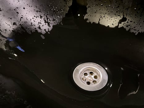 Water collects in sink with circular drain, surrounded by glossy, dark surface. Droplets cling to edges, reflecting faint light. Atmosphere textures. Details of bathroom interior, hygiene photo