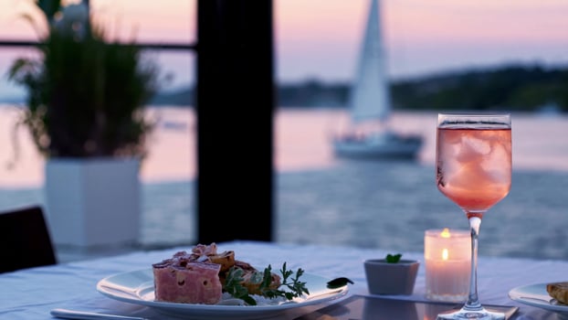 Romantic seaside dinner featuring candlelit table, rose wine, decadent dessert alongside distant sailboat silhouette during golden hour sunset lighting photo