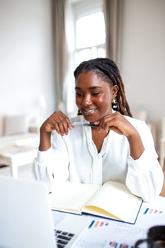 Focused young woman smiles thoughtfully while working on her laptop, pen in hand and notebook open. Charts and data hint at a productive remote work or study session. photo