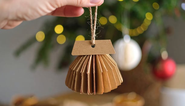Handcrafted paper ornament hanging from string, showcasing unique design with folded layers. background features festive tree adorned with colorful lights and decorations, creating warm holiday photo