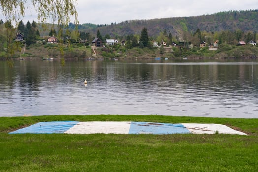 Deflated blue and white trampoline by lake in Nova Zivohost with houses and hills in background photo