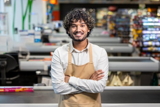 A smiling man with curly hair, wearing an apron, stands confidently with his arms crossed in a supermarket. photo