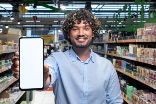 A smiling man in a grocery store holds up a blank smartphone screen, likely for advertising or app use. photo