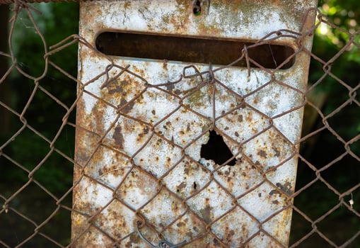 An old, weathered mailbox behind a rusty, damaged chain-link fence in an outdoor setting. photo