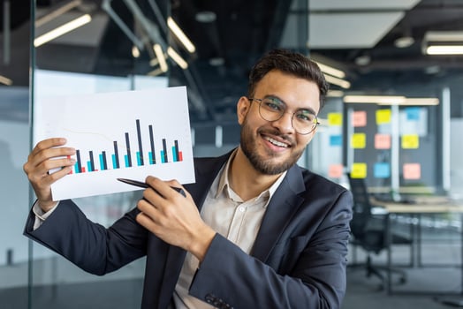 A smiling businessman in glasses points to a chart, likely in a business presentation. photo