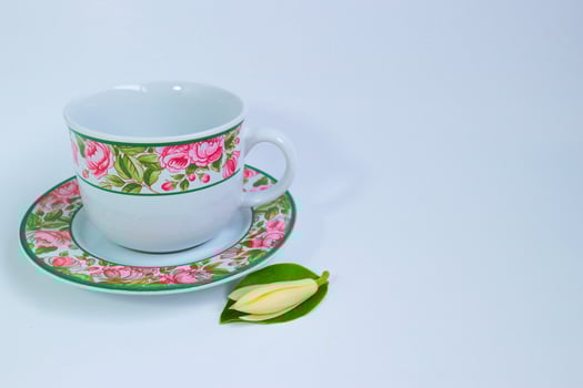 Thai ceramic cup and saucer with rose motifs, placed beside a white champa flower and leaf. Minimal studio scene with white background and right-side copy space. photo
