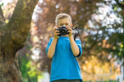 A cute boy in a blue T-shirt takes pictures with a camera in the park. Child blogger. Child photographer photo