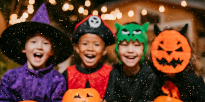 Four children in colorful costumes purple witch, pirate, green monster, and orange pumpkin smile widely while holding bright orange pumpkins under string lights. photo