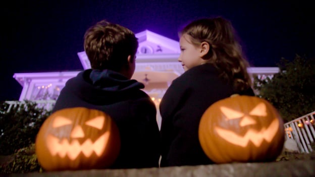 Two children sit on steps, illuminated by carved jack o lanterns with grinning faces. A grand house is softly lit in purple behind them, creating a festive mood. photo