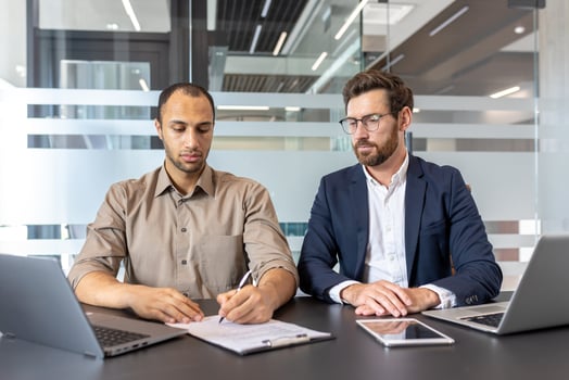 Two diverse men at a desk in an office, one signing a document while the other observes, laptops and tablet present. photo