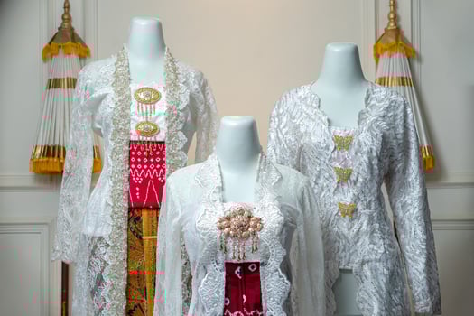 Three traditional Indonesian bridal kebayas in elegant white lace, decorated with gold brooches and red batik details, displayed on mannequins to highlight cultural wedding fashion. photo