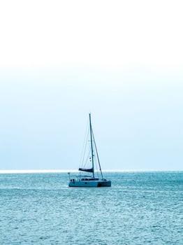 A low-angle shot captures a peaceful and expansive ocean scene. A white catamaran with its mast and rigging stands alone in the center, sailing calmly on the shimmering blue water. The horizon is a photo