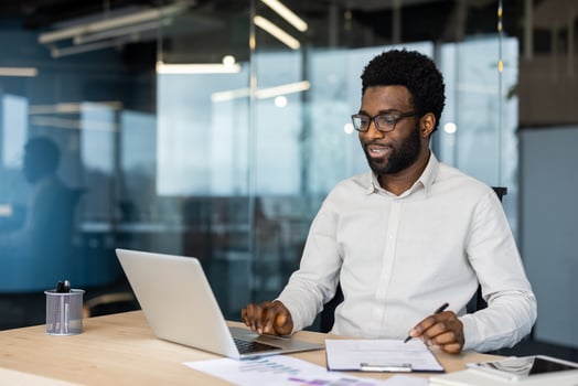 A focused individual working on a laptop at a desk in a contemporary office. The environment suggests productivity, professionalism, and a modern work setting with a hint of collaboration. photo