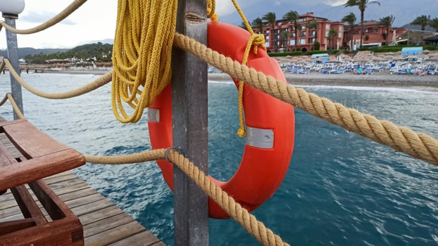 Close-up of an orange lifebuoy attached to a wooden pier with ropes, overlooking a coastal resort beach and the calm sea in the background. photo