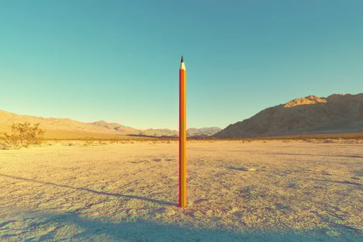 A tall orange pencil stands upright in the desolate, sunlit desert against distant mountains photo