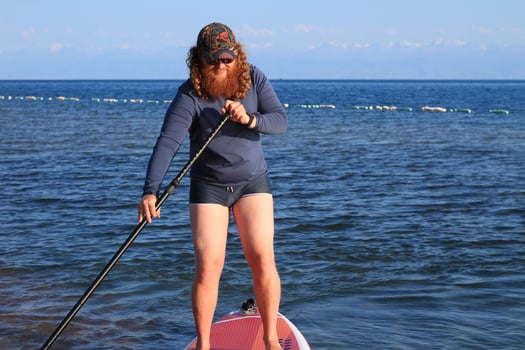 A man with a long ginger beard stands on a paddleboard in calm waters, wearing a dark long-sleeve shirt and shorts. The background features distant mountains. photo