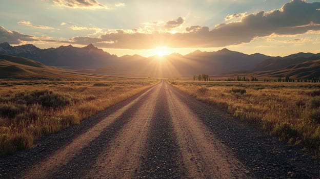 Sunlight streams above the open dirt road and dry grass with mountains in distance photo