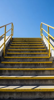 Outdoor concrete stairs with bright yellow handrails ascend towards a clear blue sky photo