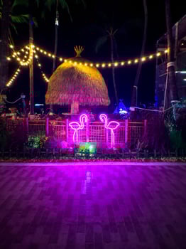 A low-angle shot captures a tropical beachside bar at night, illuminated by vibrant neon lights. Two pink neon flamingo signs stand out in the center, in front of a bamboo fence and a thatched-roof photo