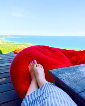 A low-angle shot captures a serene vacation moment with a woman's feet, painted with blue nail polish, resting on a bright red beanbag chair. The chair is placed on a wooden deck, with a stunning photo