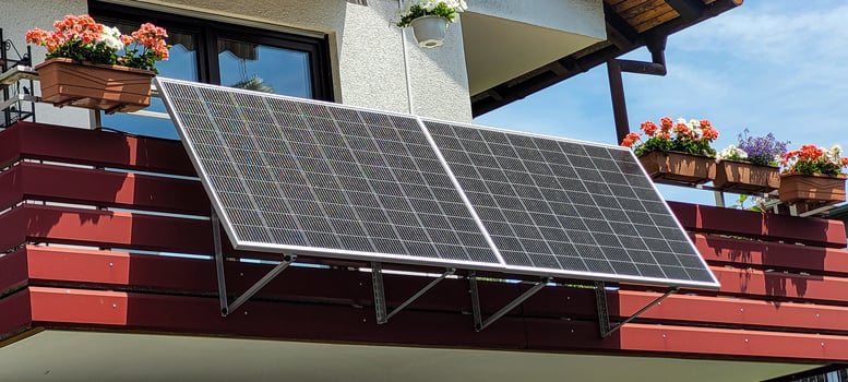 Balcony solar system, fixed at an angle of 30 degrees on the red balcony of an apartment building, surrounded by pots with pink and white geranias and other flowers of white and blue colors photo