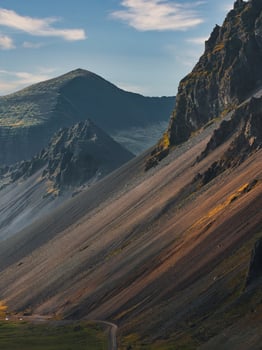 A dramatic Icelandic mountainside with steep ridges, rocky and grassy textures, a winding road below, and peaks under a clear sky with scattered clouds. photo