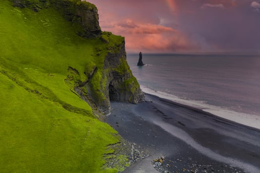 Iceland's black sand beach features green cliffs, a sea stack, and a pink orange sky at sunset. A faint rainbow adds to the dramatic coastal scene. photo
