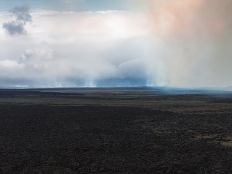 Dark volcanic terrain with cooled lava fields stretches into the distance under low clouds and mist, with hints of orange and blue in the sky. photo