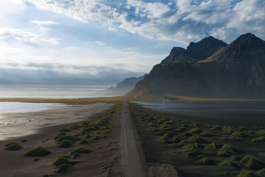 Aerial view of Iceland's Stokknes Peninsula showing a dirt road, black sand, green mounds, jagged mountains, and a reflective body of water. photo