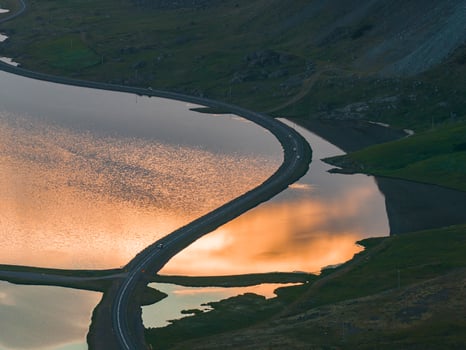 A serene aerial view of a winding road dividing a reflective lagoon during sunset, with orange skies mirrored in the water and rocky green terrain. photo
