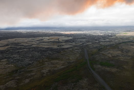 A winding road traverses a barren, rocky terrain with moss patches and sparse vegetation under a dramatic sky with thick clouds and orange glow. photo