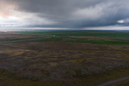 Vast Icelandic terrain with rocky foreground, green fields, and roads. Overcast sky casts shadows, with a distant settlement near the horizon. photo