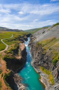Studlagil Canyon features basalt columns, a turquoise river, greenery, rocky terrain, a dirt path, a winding road, and a clear blue sky with clouds. photo