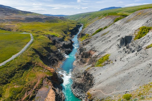 Studlagil Canyon in Iceland features a turquoise river, steep rocky cliffs, green vegetation, a dirt road, and a clear blue sky with distant hills. photo