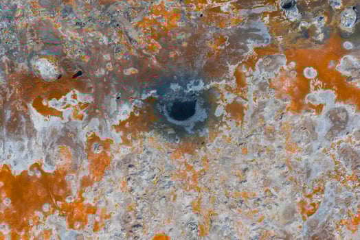 Aerial image of a geothermal area in Iceland featuring vibrant orange, white, and gray terrain, a dark mud pot, mineral deposits, and visible steam. photo