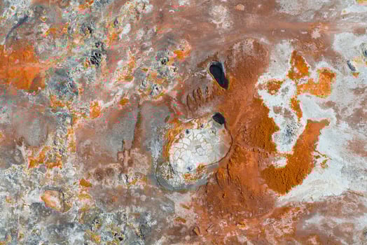Geothermal terrain in Iceland with vibrant orange, red, and white hues, featuring two dark oval pools surrounded by cracked, pale surfaces. photo