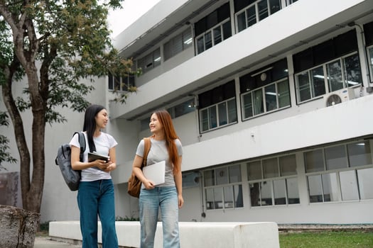Two friendly students walking together on campus, carrying laptops and backpacks, sharing a light moment photo