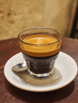 Shot of espresso with layer of crema on top served in small glass cup on white saucer with metal spoon, placed on wooden table in cozy setting photo