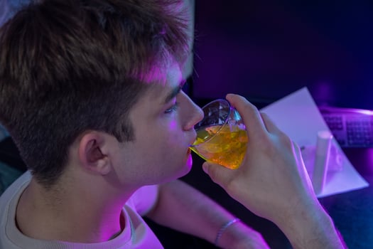 Young man drinking medicine dissolved in water or energy drink at night, working on computer, illuminated by bright pink neon lights, photo