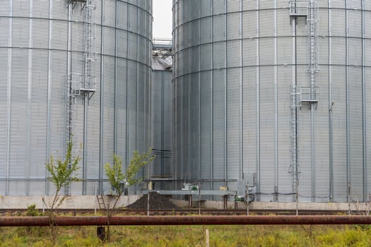 Two tall metal silos are positioned side by side at a rural farm, with green grass and a rusty pipeline in the foreground under a cloudy sky photo