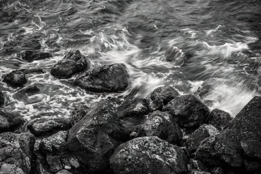 A high-angle, black and white long exposure shot of the sea. Water blurs as it washes over a rocky shoreline, creating a moody and dynamic texture with a monochromatic tone. photo