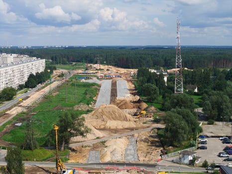 Large scale road construction on Riga to Jelgava road with excavators and cranes, bordered by a residential building, a communication tower, green spaces, and a forested area. photo
