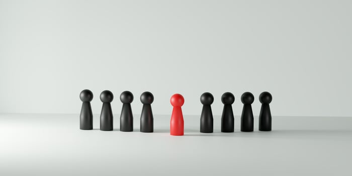 Individuality, leadership, being different, and standing out in a team or group. A single red doll prominently positioned among a line of black dolls on a vibrant white background. photo
